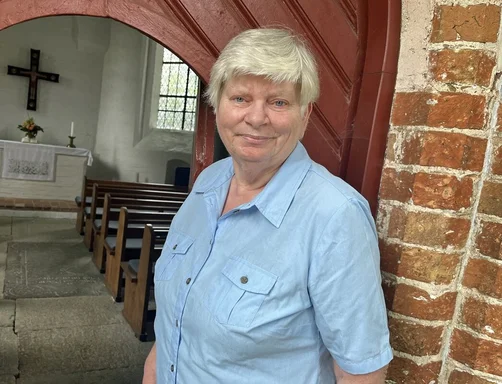 Eine Frau mit kurzen grauen Haaren steht vor dem Eingang einer Kapelle.