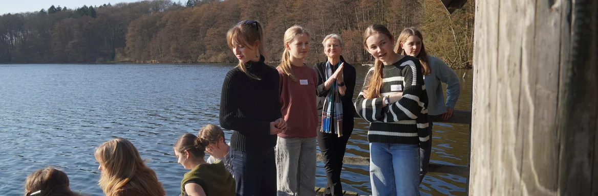 Junge Menschen und eine Frau stehen und sitzen auf einem Steg an einem See.