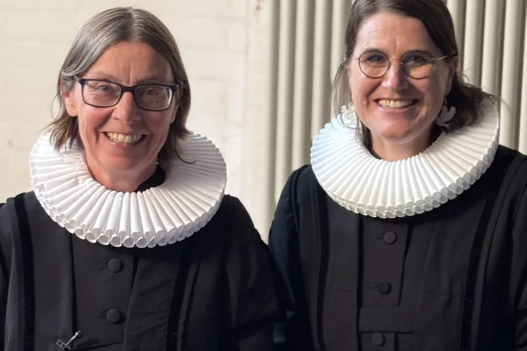 Twei Frauen stehen im Talar und mit Halskrause nebeneinander und lächeln in die Kamera.