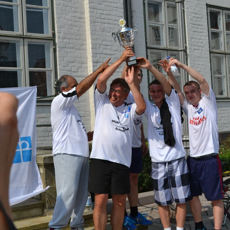 Mehrere Fünf Personen mit kurzen Hosen und weißen T-Shirts halten gemeinsam einen Pokal in die Luft