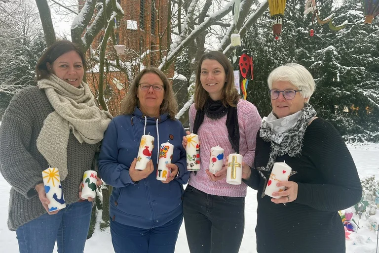 Vier Frauen stehen auf einem verschneiten Friedhof und halten bunte Kerzen in den Händen