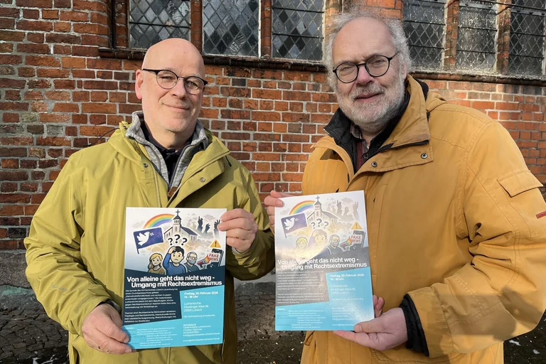 Zwei Männer in hellen Jacken halten zwei Plakate in die Kamera.