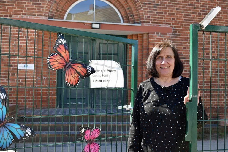 Eine ältere Frau mit dunklen Haaren steht an einer Pforte. 