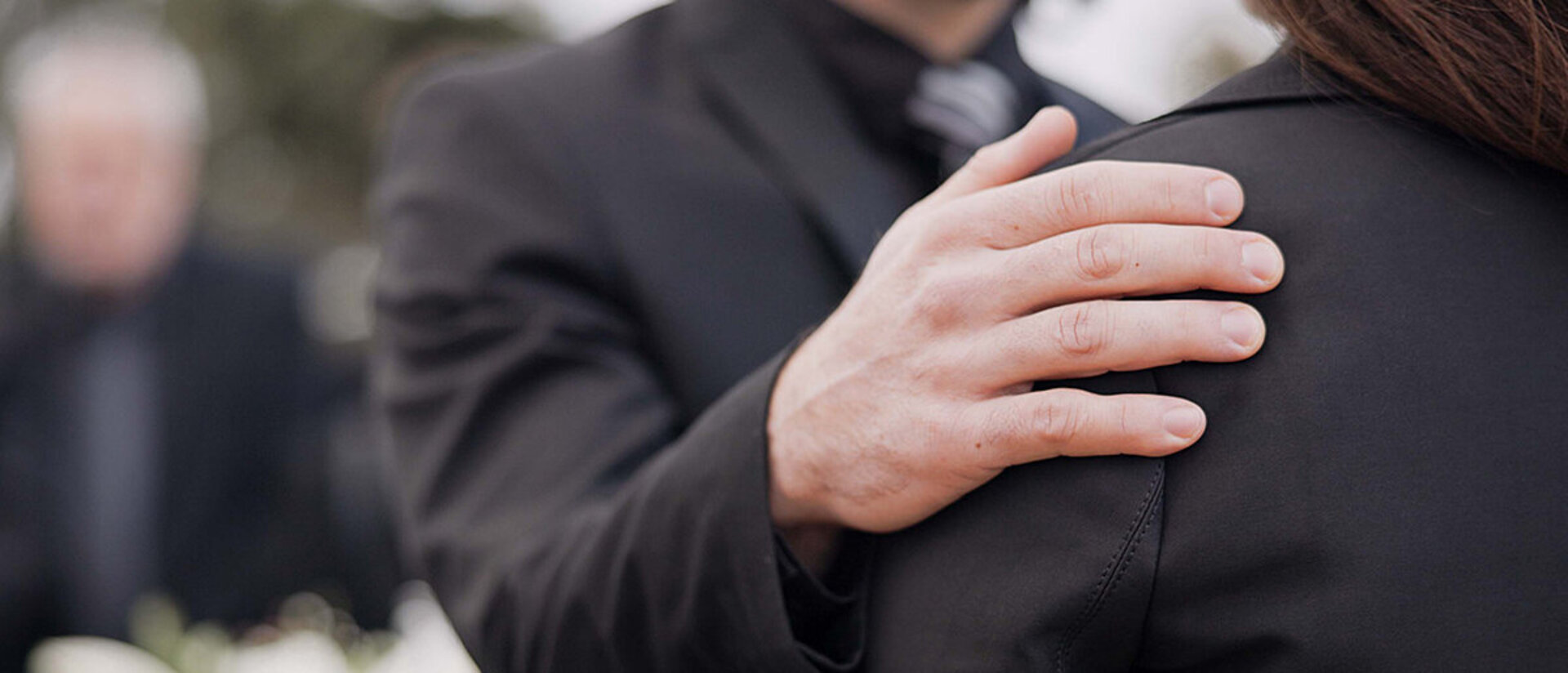 Im Hintergrund verschwommen sieht man einen weißen Blumenstrauß und eine Person mit dunklem Anzug. Im Vordergrund scharf abgebildet hält eine Mann, dessen Kopf man nicht sehen kann, seine rechte Hand tröstend auf der Schulter einer Frau in schwarzer Kleidung und dunklen Haaren. Auch ihr Gesicht kann man nicht sehen. Der Mann trägt ebenfalls schwarze Kleidung: Ein schwarzes Sakko und ein dunkles Hemd mit dunkler Krawatte.