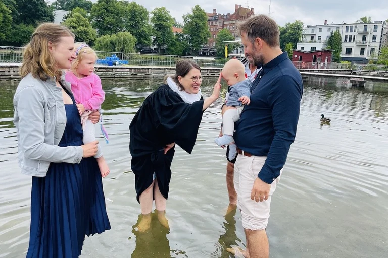 Nach Töchterchen Eva (vor zwei Jahren) lassen Marlene und Tobias Reinberger Sohnemann Johan von Pastorin Margrit Wegner taufen. Sie alle stehen im Wasser, die Pastorin lächelt und tauft Johan.