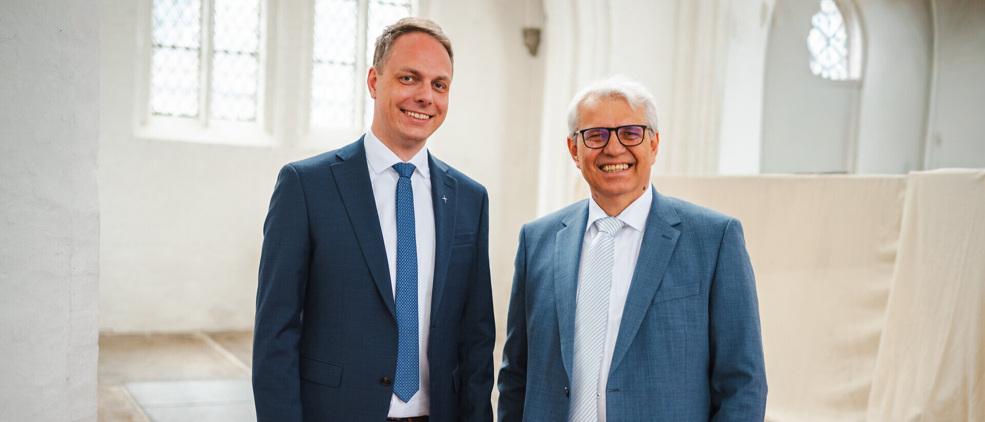 Zwei Männer stehen nebeneinander: Der linke mit weißem Hemd, hellblauer Krawatte und dunkelblauem Anzug, der rechte trägt ein weißes Hemd und ein hellblaues Sako. Im Hintergrund eine leere Kirche mit weißen Wänden und Boden aus Ziegelsteinen.