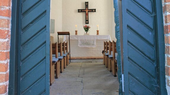 Eine offene blaue Kirchentür gibt den Blick frei zu einem Kirchenaltar auf dem zwei große, weiße, brennende Kerzen zu sehen sind. Zwischen den Kerzen steht eine runde Glasvase mit Blumen, darüber hängt ein Altarkreuz. Im vordergrund sind braune Holz-Kirchenbänke mit blauen Sitzauflagen zu sehen.