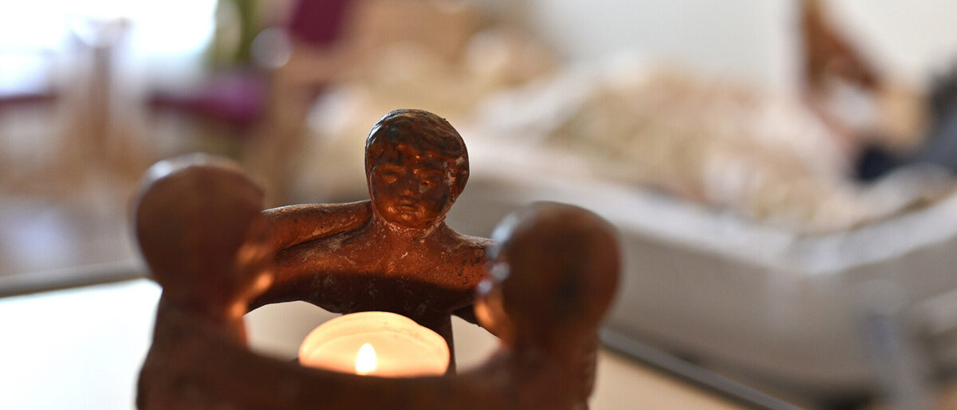 Im Hintergrund verschwommen liegt eine Person in einem Bett. Im Vordergrund ist ein Teelicht zu sehen, welches von drei Figuren umschlossen wird.