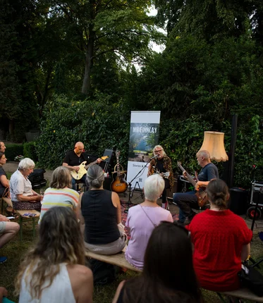 Menschen sitzen im Grünen und hören einem Konzert zu. 