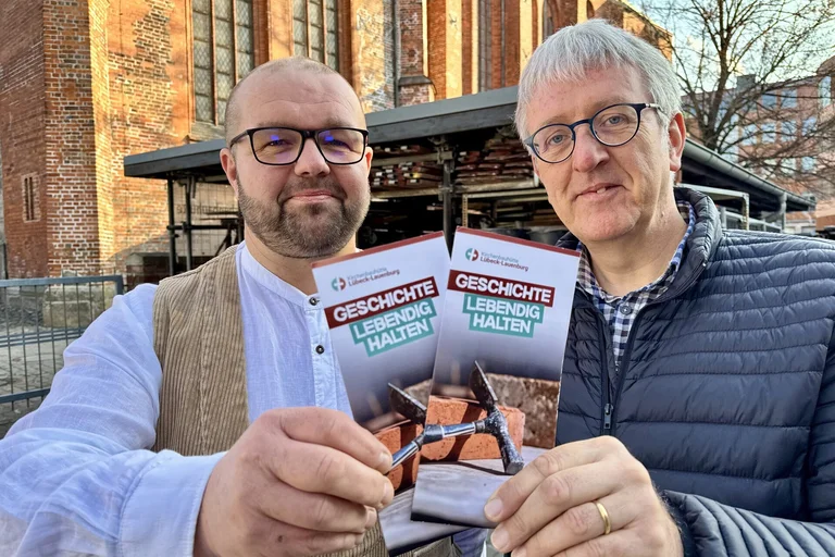 Zwei Männer stehen vor einer Kirche und halten einen Flyer in die Kamera