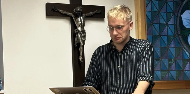 Ein Mann steht an einem Pult vor einem Kreuz an der Wand.