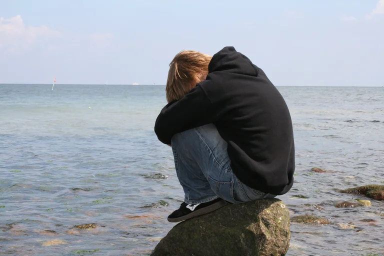 Eine Person in Jeans und dunklem Pullover und Kaputze sitzt am Meer auf einem Stein. Die Person hat kurze Haare, die Beine angezogen, die Arme umgeben die Beine, der Kopf ist gesenkt. Das Gesicht nicht sichtbar in den Armen versteckt.