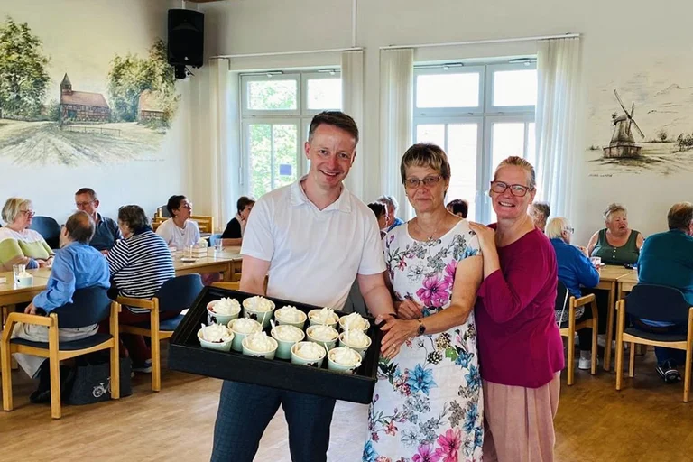 Ein Mann und zwei Frauen stehen in einem Speiseraum und zeigen ein Tablett mit Eisbechern.