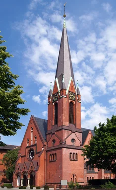 Turm in Mitte über Eck, links Langhaus-Giebel und rechts Südschiff-Giebel, beidseitig grüne Bäume