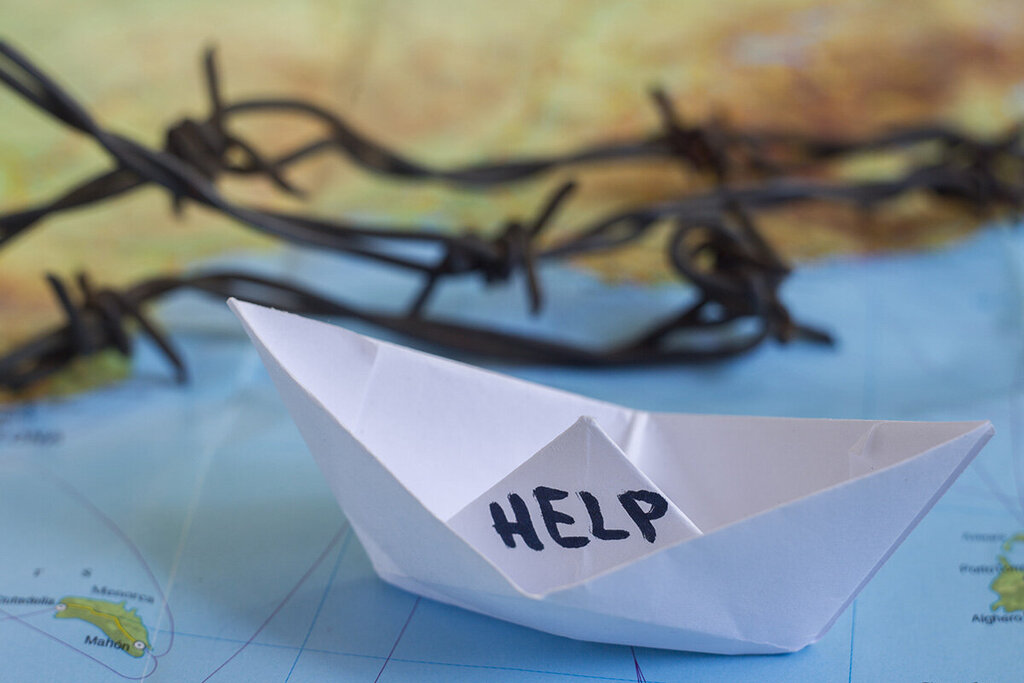 Ein Papierschiffchen mit der Aufschrift "Help" steht auf einer Wasserfläche einer Landkarte. Im Hintergrund ist eine Stacheldraht zu sehen 