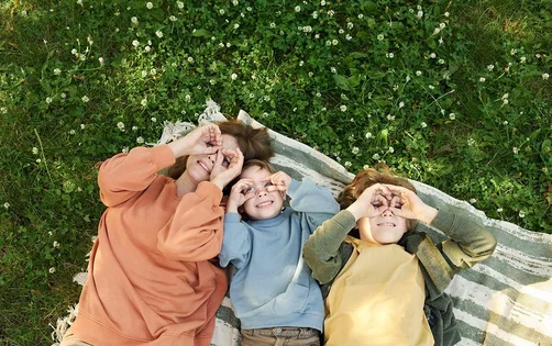 Draufsicht auf eine Frau und zwei Jungen, die zusammen auf einer Picknickdecke liegen und lächelnde Gesichter machen - sie halten die Finger zu einer Brille an die Augen.