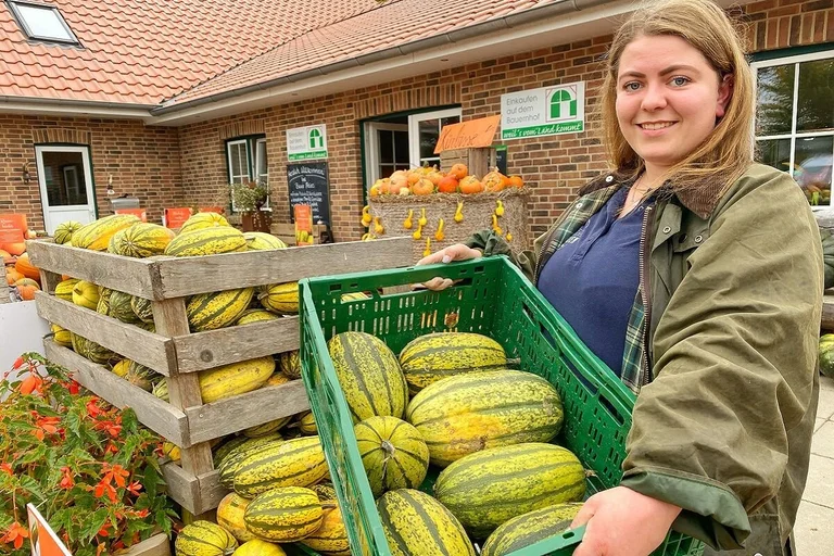 Junge Frau steht mit einer Kiste mit Kürbissen vor einem Hofladen und lächelt in die Kamera. 