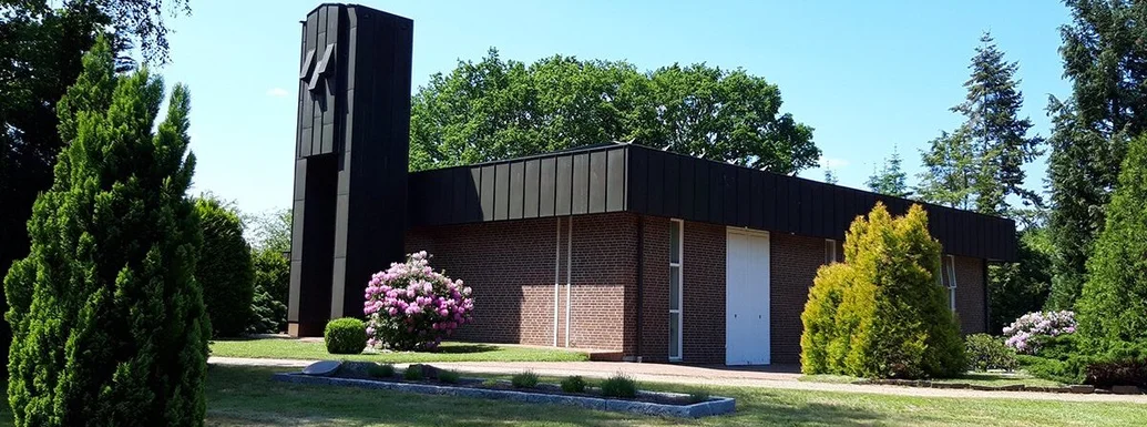 Kapelle Waldfriedhof steht im Hintergrund bei strahlend blauem Himmel. Links und rechts sind Bäume und Sträucher. Im Vordergrund Rasen und links ein kleiner grüner Baum