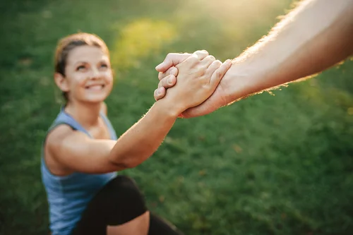 Eine Frau sitzt auf dem Rasen und hält die Hand einer Person von der nicht mehr zu sehen ist. Es scheint die Sonne, die man aber nicht sieht. Die Frau lächelt.
