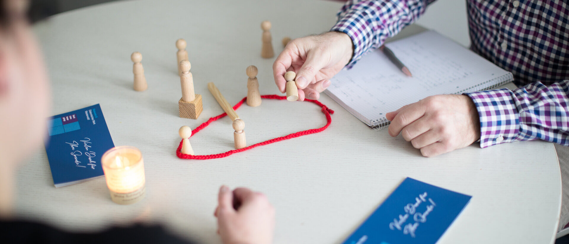Zwei Personen sitzen an einem hellen runden Tisch, darauf stehen kleine Holzfiguren teils in einem roten Ring.