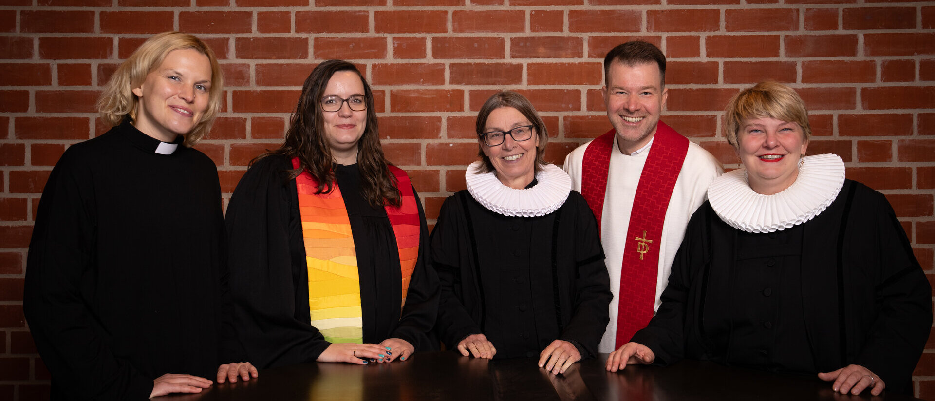 Vier Frauen und ein Mann in Pastorengewändern stehen vor einer Backsteinwand an einem dunklen großen Tisch.