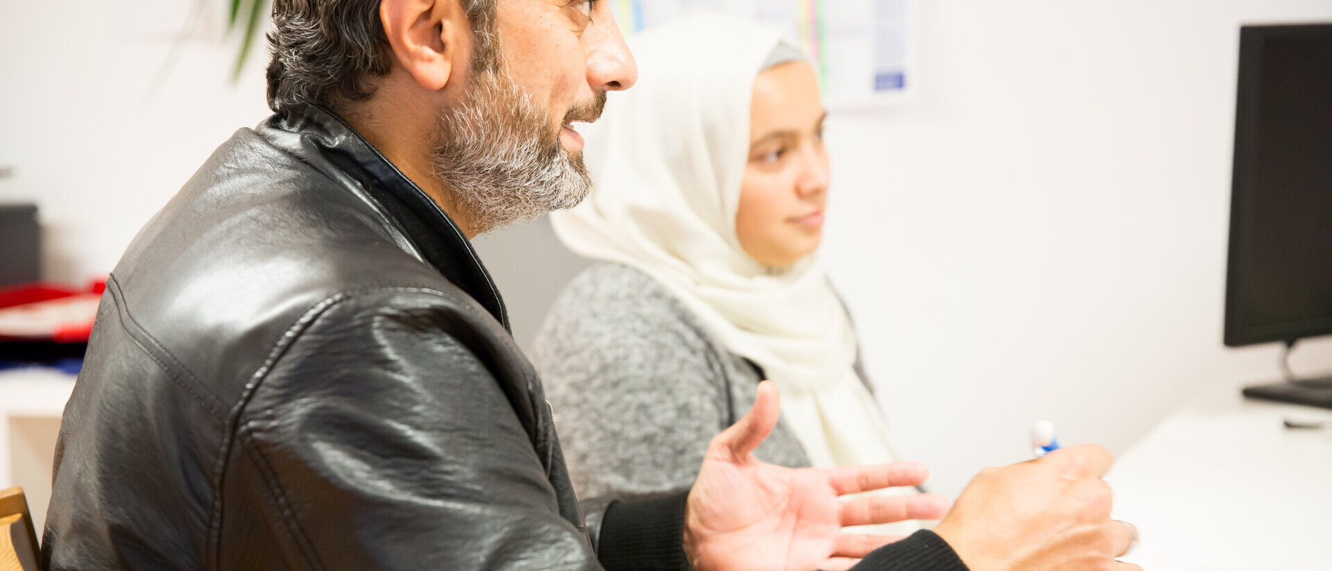 Eine Frau und ein Mann mit Migrationshintergrund sitzen vor einem Tisch. Die Frau trägt ein helles Kopftuch